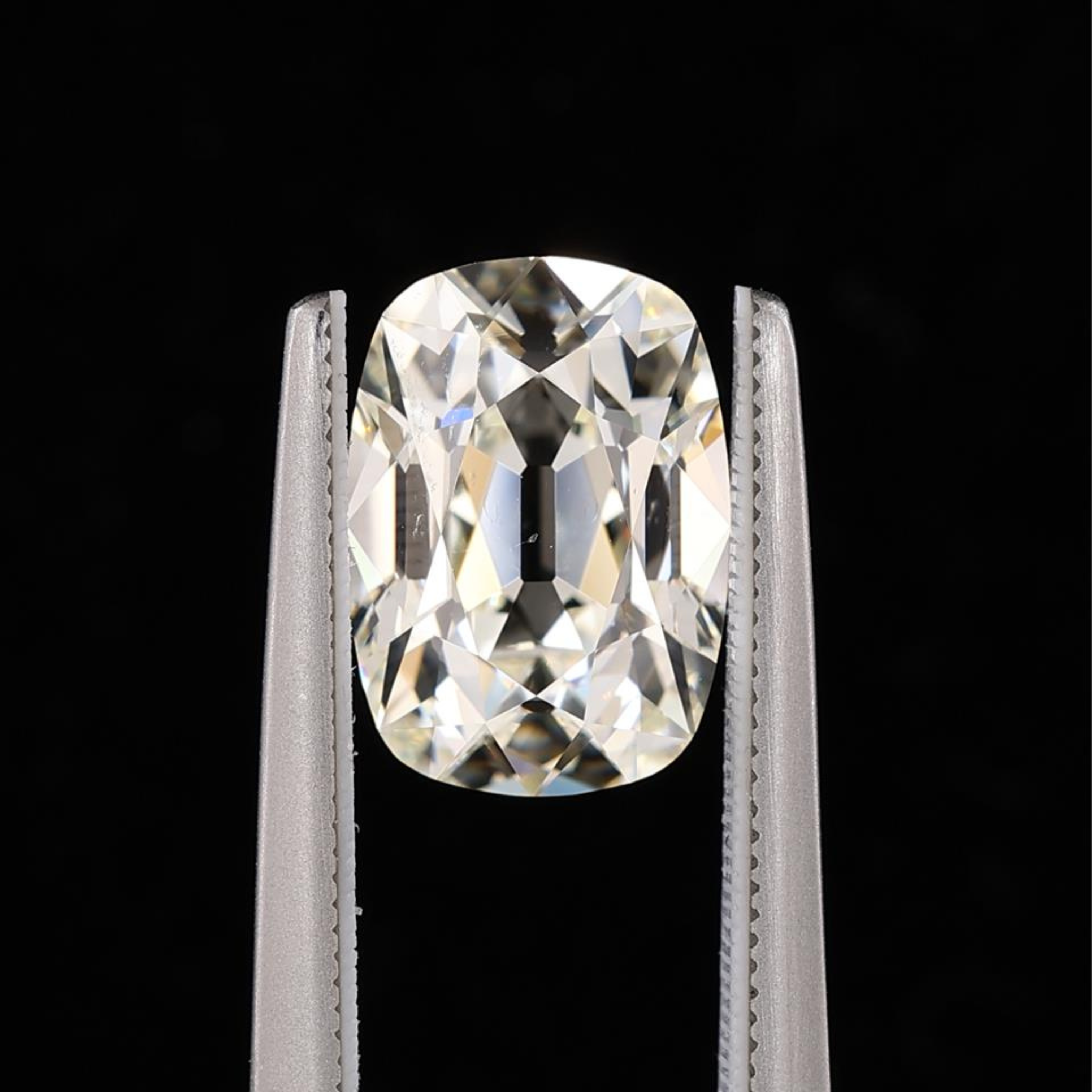 A cushion-cut brilliant gemstone held in the jaws of silver tweezers against a solid black background.