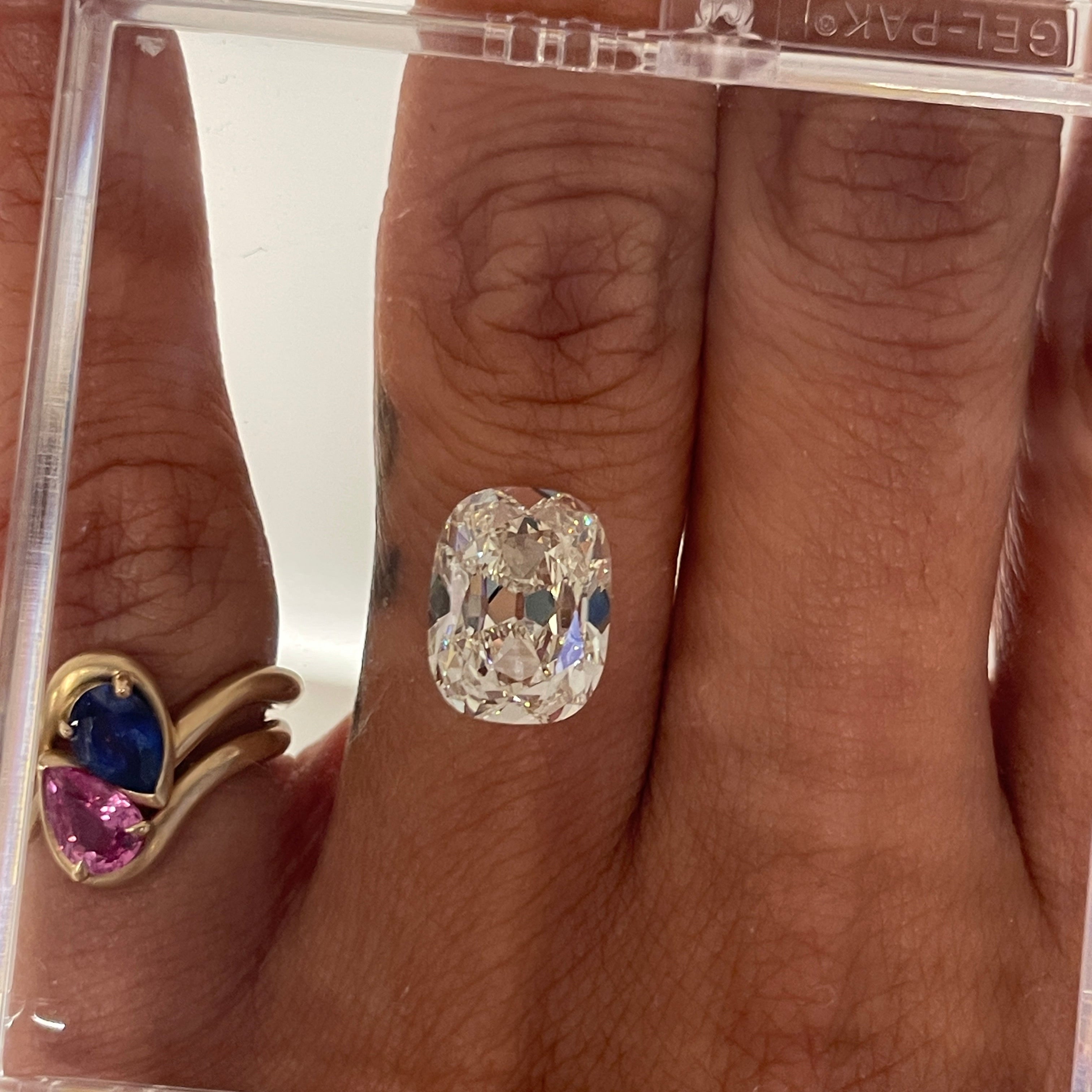 Close-up of a hand wearing a cushion diamond with a clear plastic display case. BB thelma Blue and Pink Sapphire Stacked on the pinky finger.