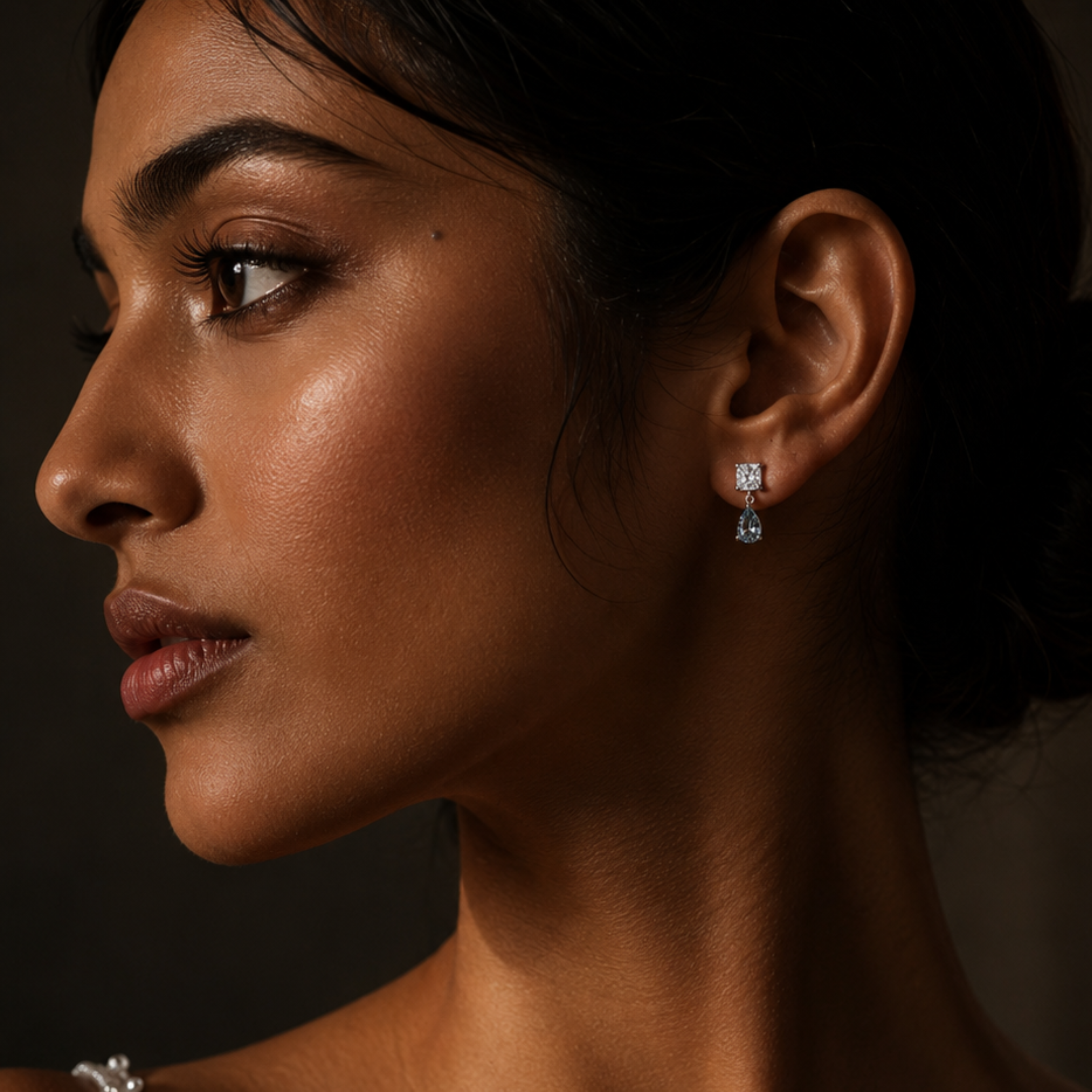Close-up side profile of a woman wearing a small silver-toned dangle earring with a clear gemstone, against a dark background.