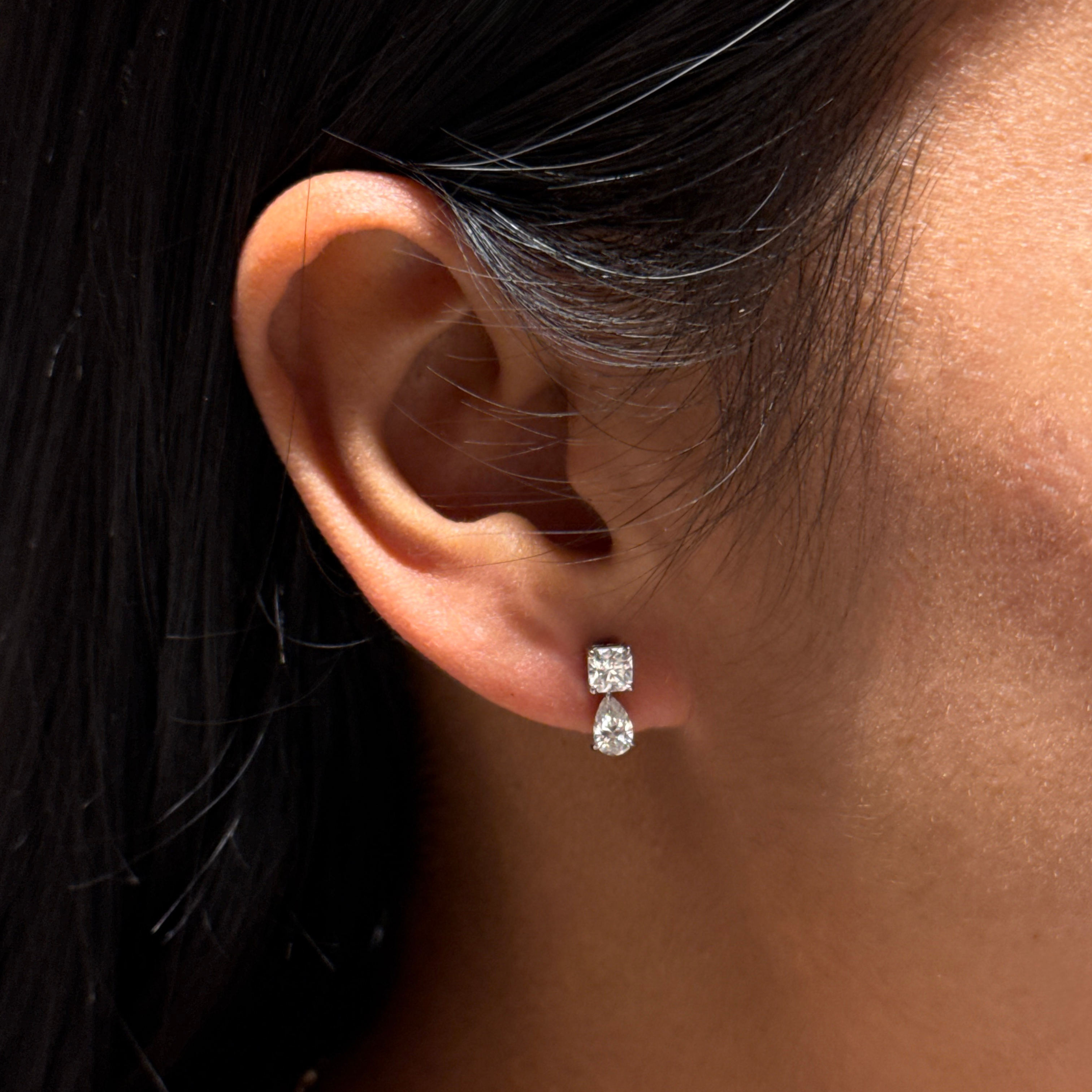 A close-up of a person's ear wearing a pair of small square-cut clear stud earrings with a platinum setting.