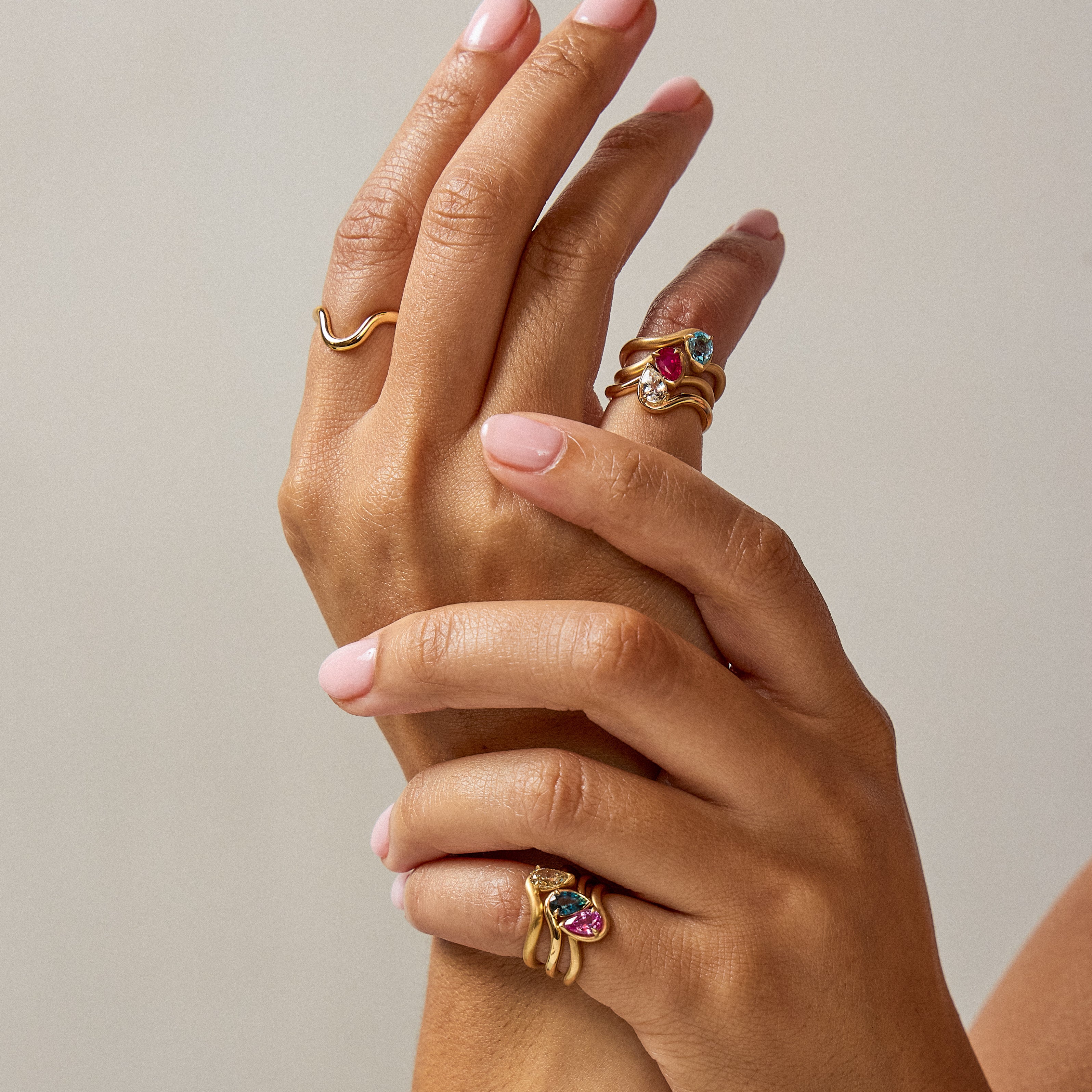 Hand model wearing BB Thelma pinky rings in pink sapphire, blue sapphire, brown diamond all in 14k yellow gold on left hand pinky and BB Thelma pinky rings in blue swiss topaz, ruby, and diamond in 14k yellow gold on right hand pinky in front of a grey background.  
