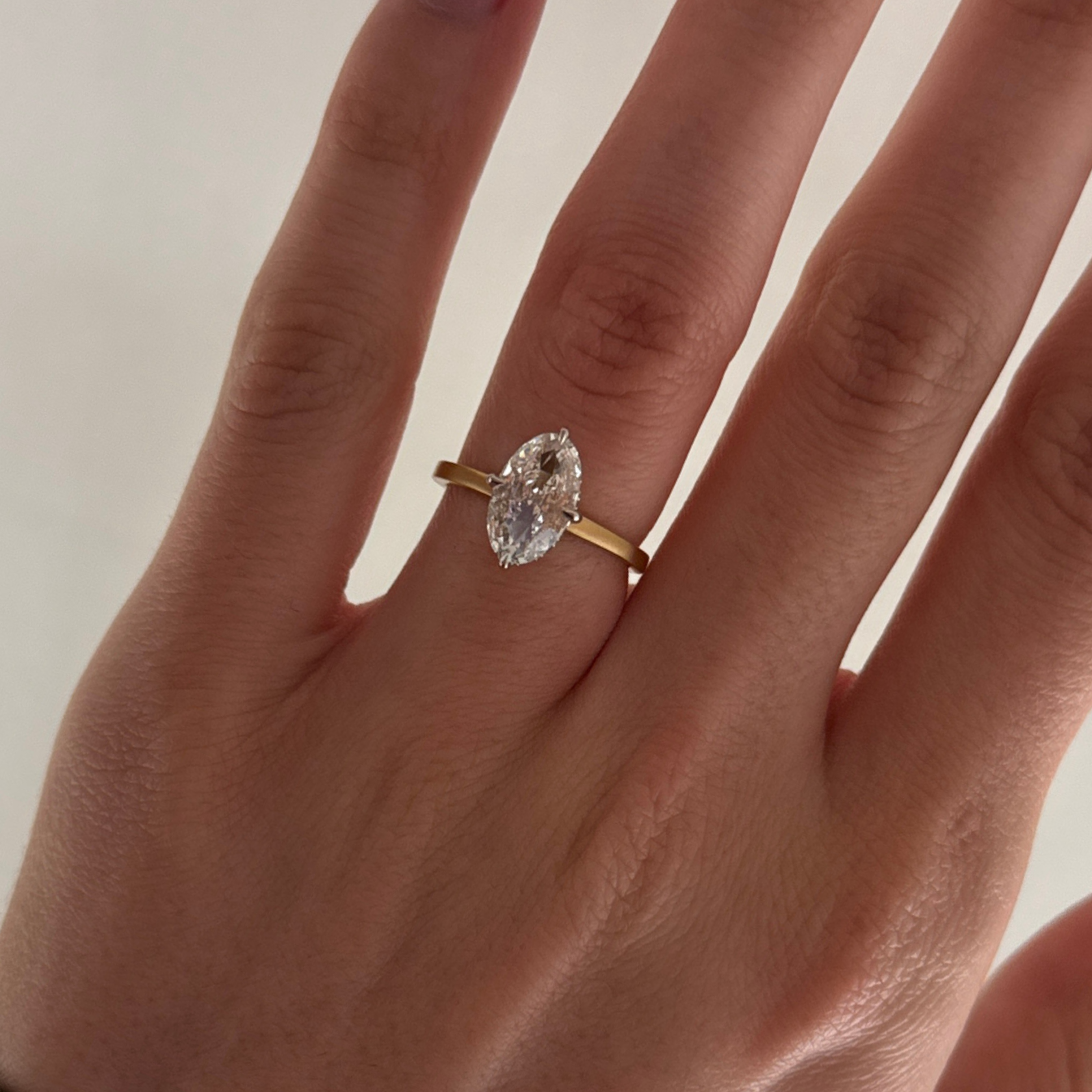 A gold ring with a Moval-cut clear gemstone worn on a finger, shown in a close-up shot of a hand against a light neutral background.