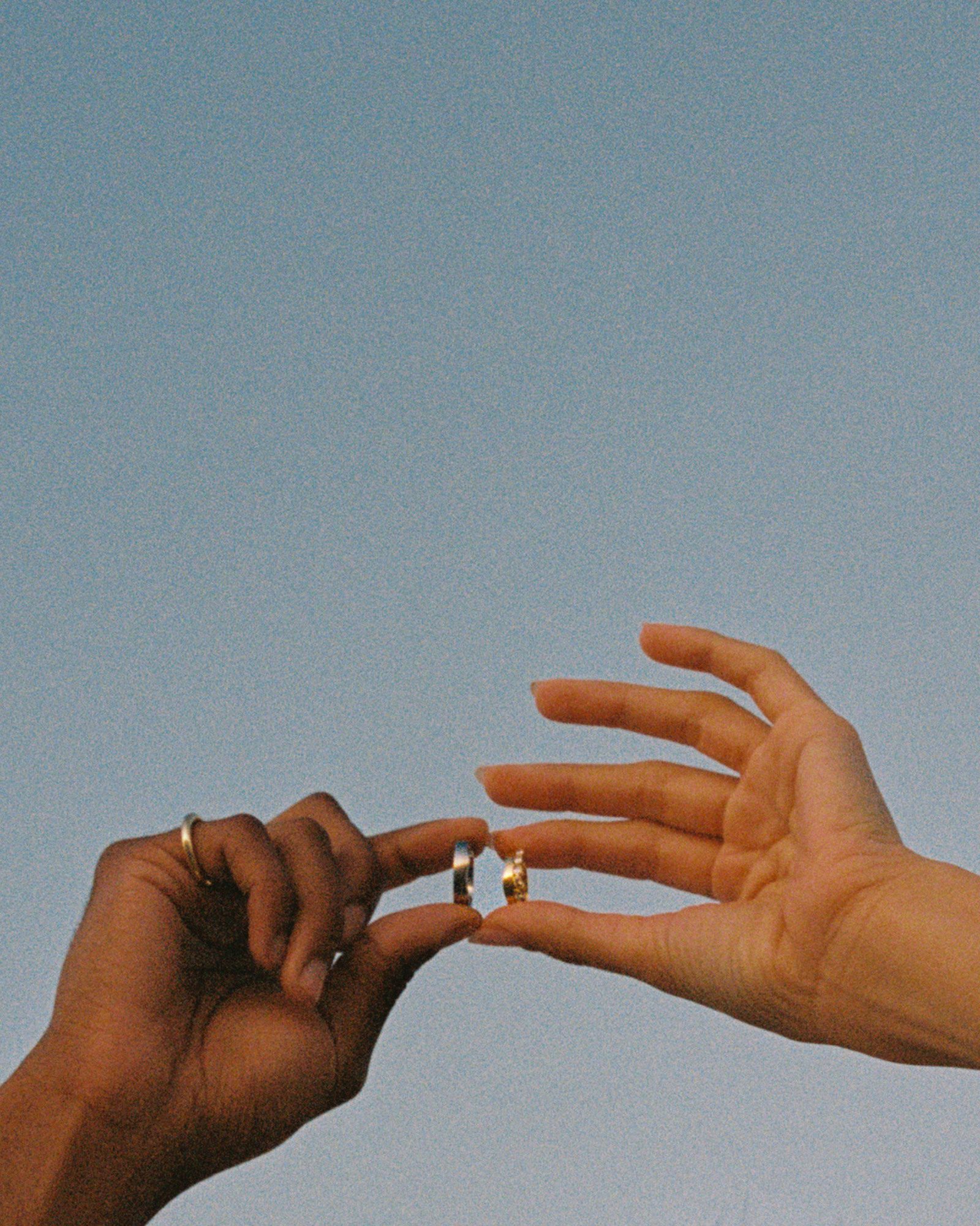 Two hands reaching toward each other against a pale blue sky, with a small gold ring held between the fingers.