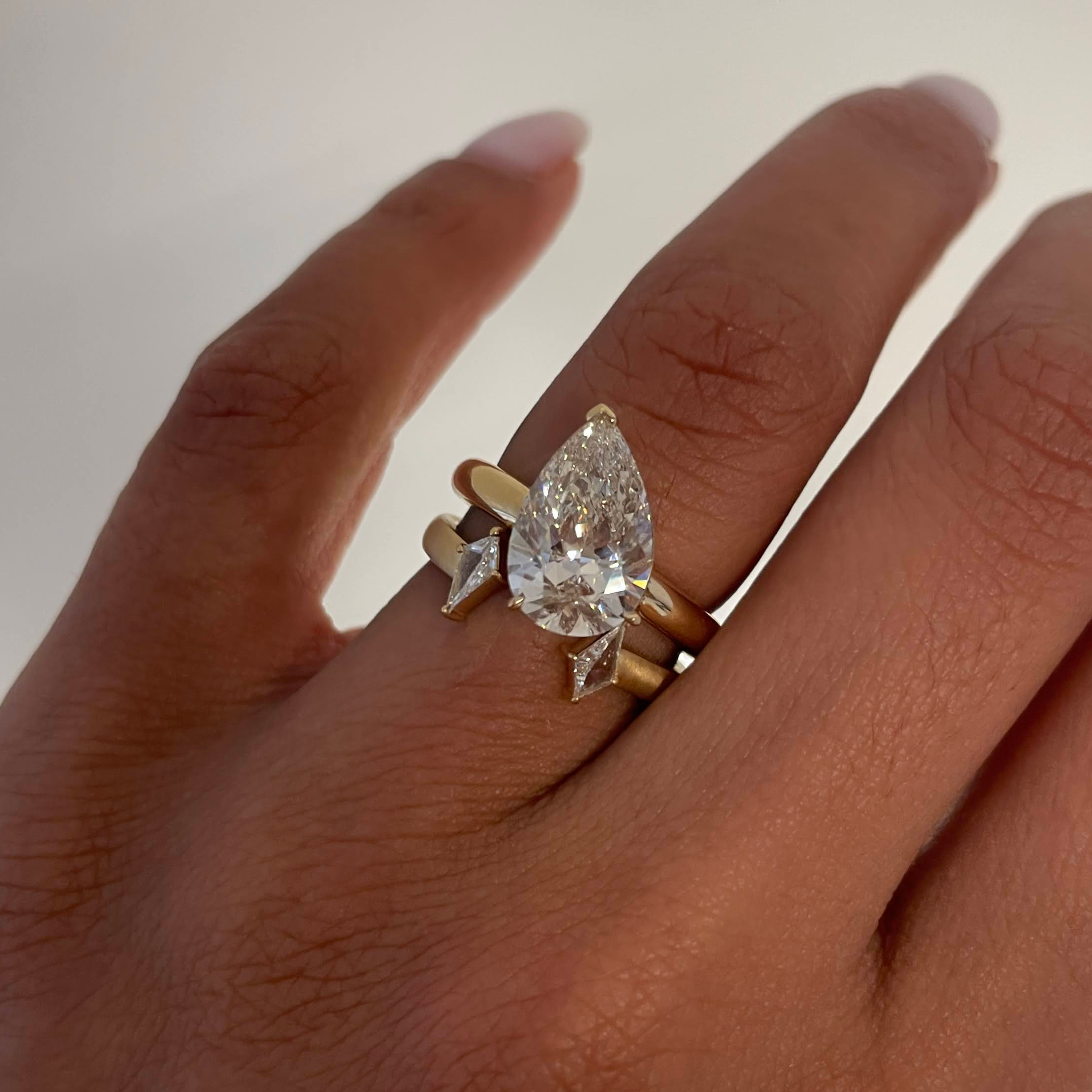 A close-up of a hand wearing a gold ring with a large pear-cut clear gemstone and smaller kite-shaped clear stones on a plain light background.