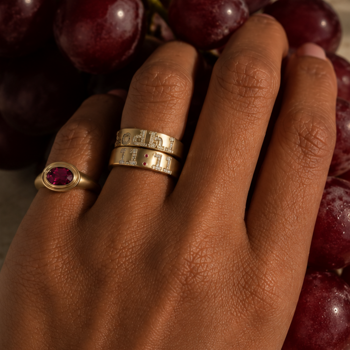 A close-up of a hand wearing two gold rings, one with a round rhodolite garnet gemstone and the other with engraved numerals, resting against a cluster of dark red grapes.
