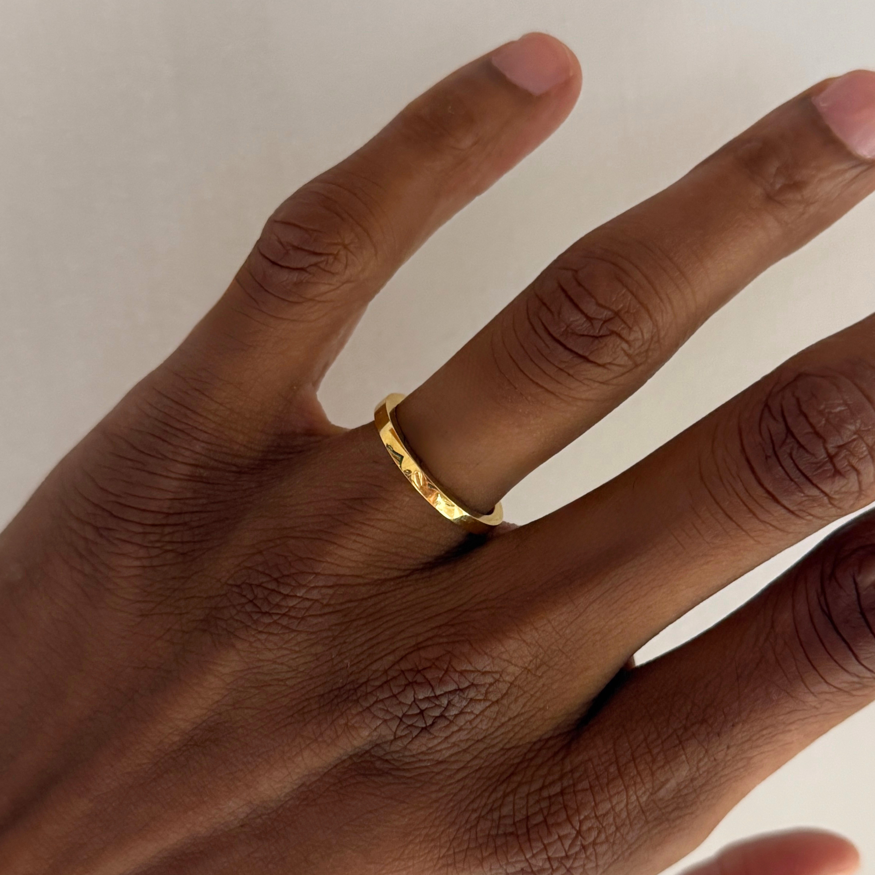 A close-up of a hand wearing a thin, textured yellow gold band ring on one of the fingers, set against a plain light background.