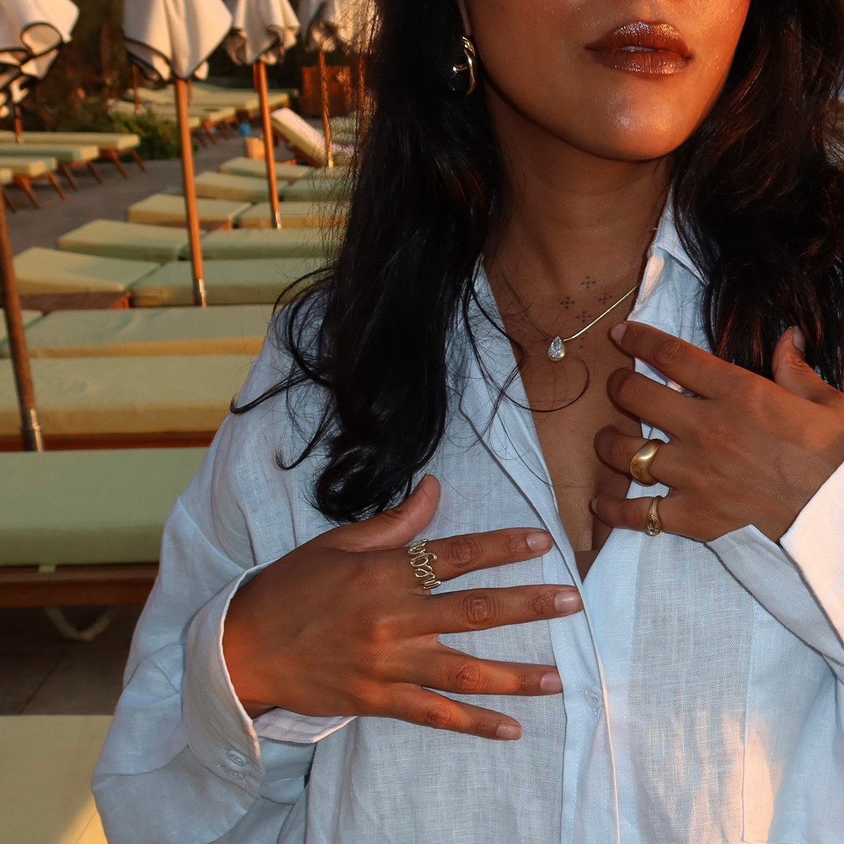 Woman wearing a name ring, pinky rings and a necklace, overlooking a sunset offscreen.