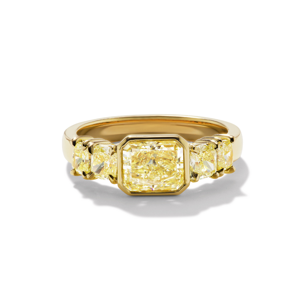 Quinn Engagement ring, A bezel set radiant cut yellow diamond ring with cushion-cut side stones in yellow gold.
